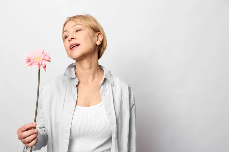 woman holding pink flower, senior portrait with calm expression, eyes closed, soft studio light on gray background, spring tenderness, wellness concept