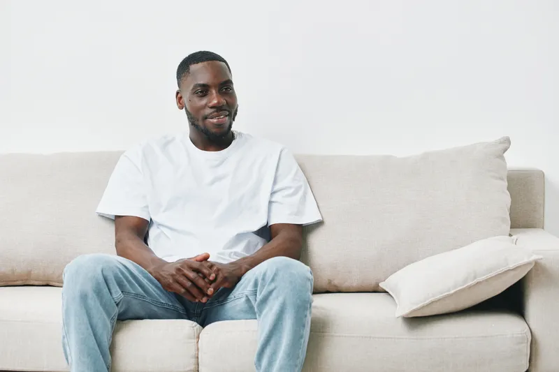 Smiling young African American man sitting comfortably on a beige couch, dressed in a casual white t shirt and blue jeans, showcasing relaxed emotions in a modern living space