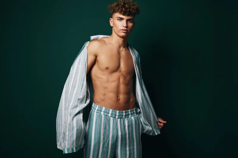 Young man with curly hair posing shirtless in striped pajama pants and open shirt against dark background. Fashion, casual style and confident expression in studio lighting.