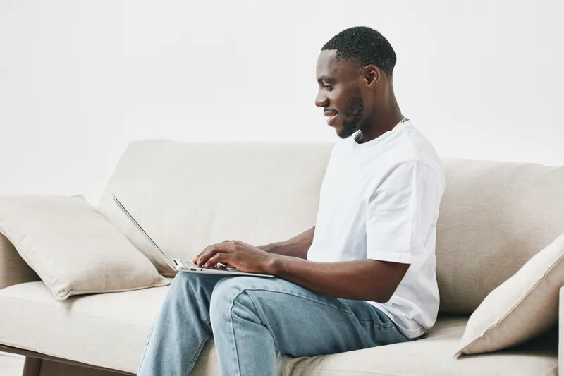 Smiling African American man using a laptop on a comfortable sofa in a modern living room, showcasing a relaxed and casual home environment