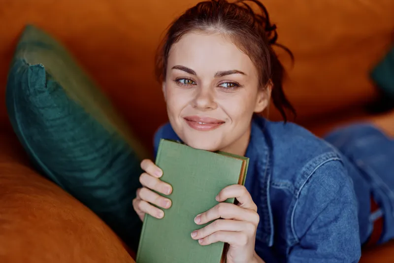 Счастливая женщина расслабляется на диване с книгой в руках, улыбаясь от удовольствия и наслаждения