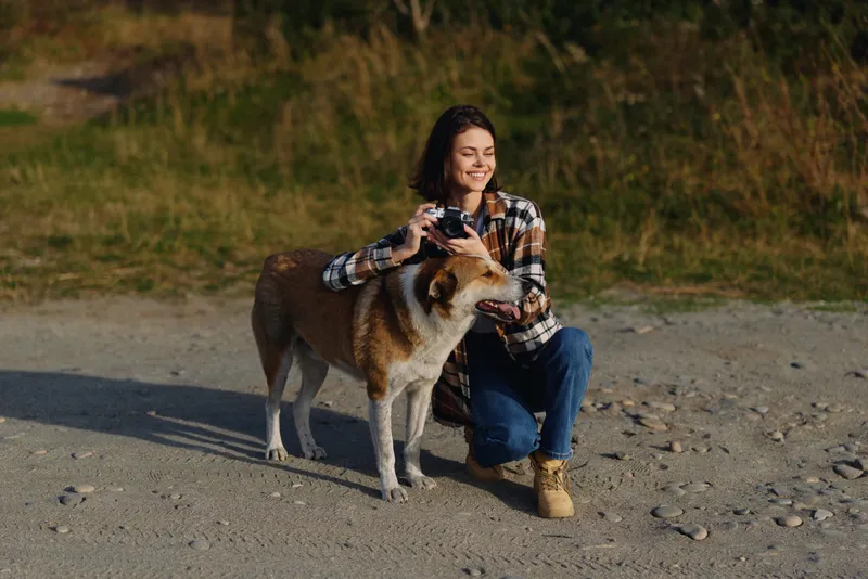Woman crouching beside a dog on a gravel path, holding a camera, in a natural outdoor setting with grass and hills, capturing candid companionship and scenic moments for documentary style photography