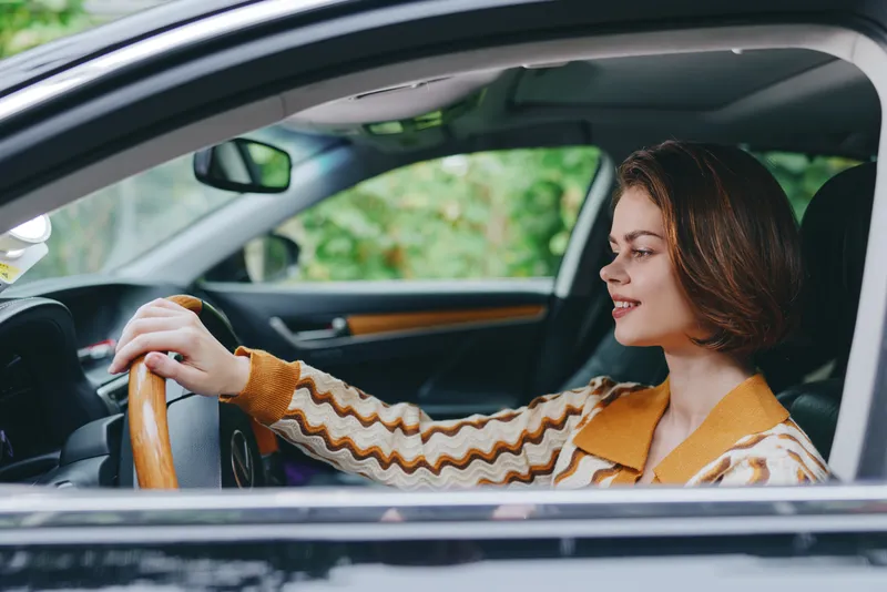 A woman driver sits behind the wheel in a car, smiling and gripping a wooden steering wheel, captured in a warm, candid moment.