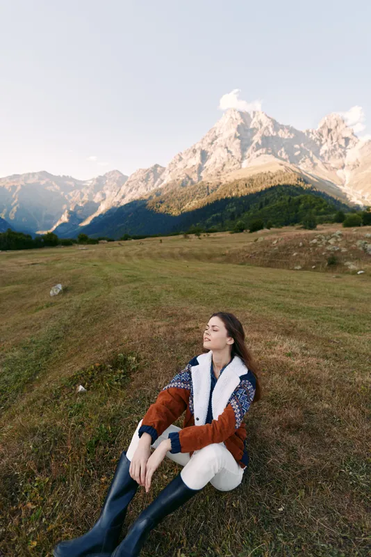 woman sitting on meadow in mountain landscape, nature scene with relaxed pose and closed eyes, outdoors lifestyle with warm jacket and boots enjoying fresh air and peaceful view