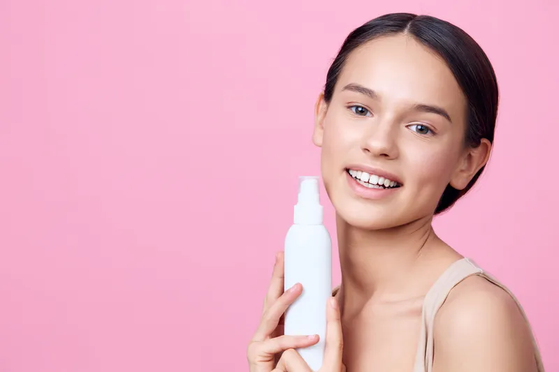 Smiling young woman holding a skincare spray against a pastel pink background, emphasizing natural beauty and healthy skincare routine