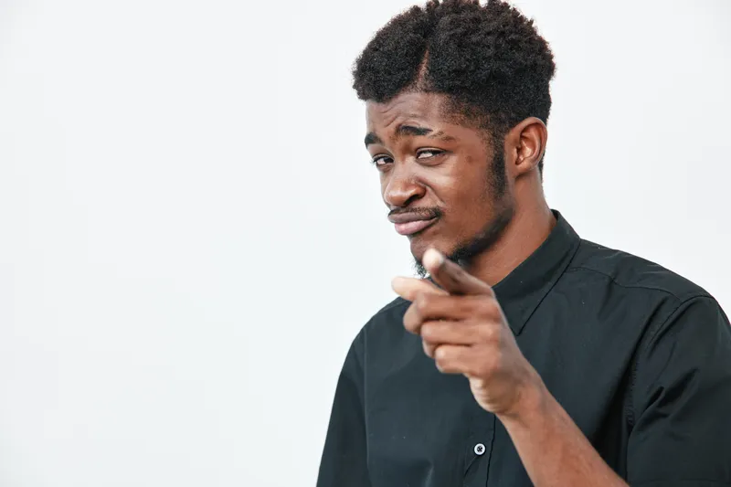 African American man with dark skin and curly hair wearing black shirt pointing finger with confident expression on white background. Portrait, gesture and casual style.