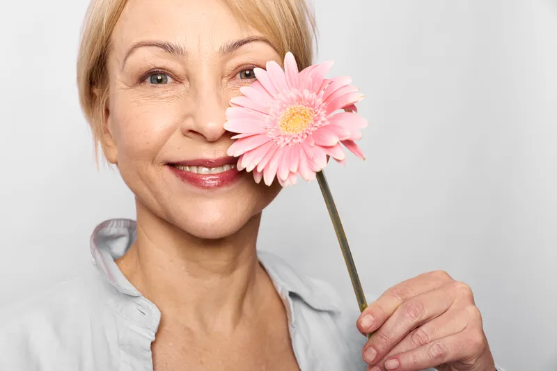 Woman portrait of a senior smiling with a pink flower near her cheek, holding the stem with fingers against a white background, spring beauty concept, soft mood