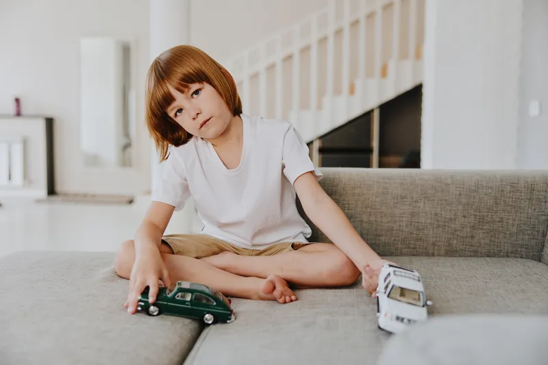 Thoughtful boy playing with toy cars on a cozy sofa, showcasing creativity and imagination in a bright, modern living room environment.