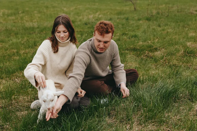 A couple outdoors in cozy sweaters feed a white goat kid in a grassy field, capturing a warm lifestyle moment of bonding, care, and nature with a friendly farm animal.