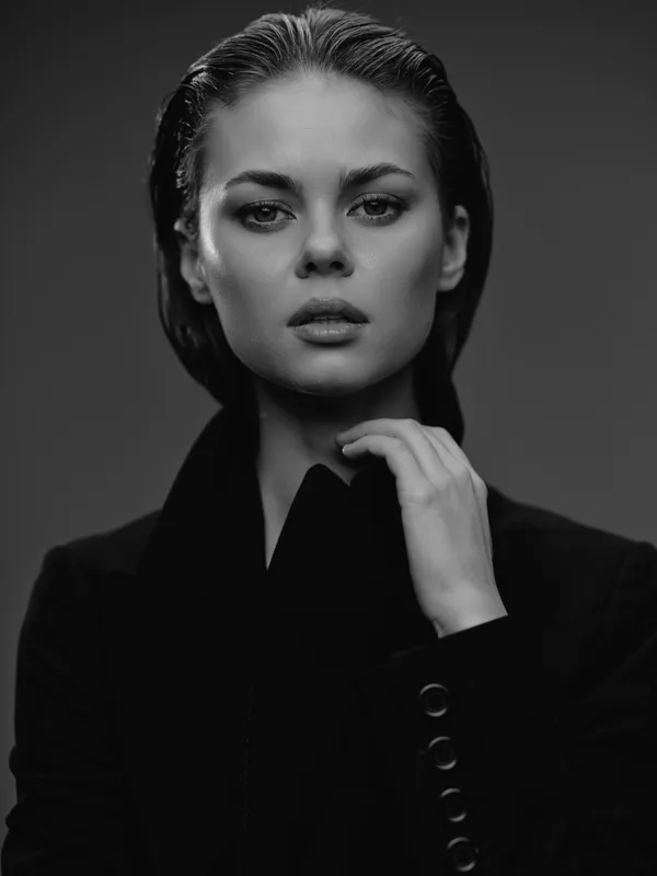 Black and white portrait of a serious woman with wet hair wearing a dark blazer, posing confidently with hand near neck, minimalistic background.