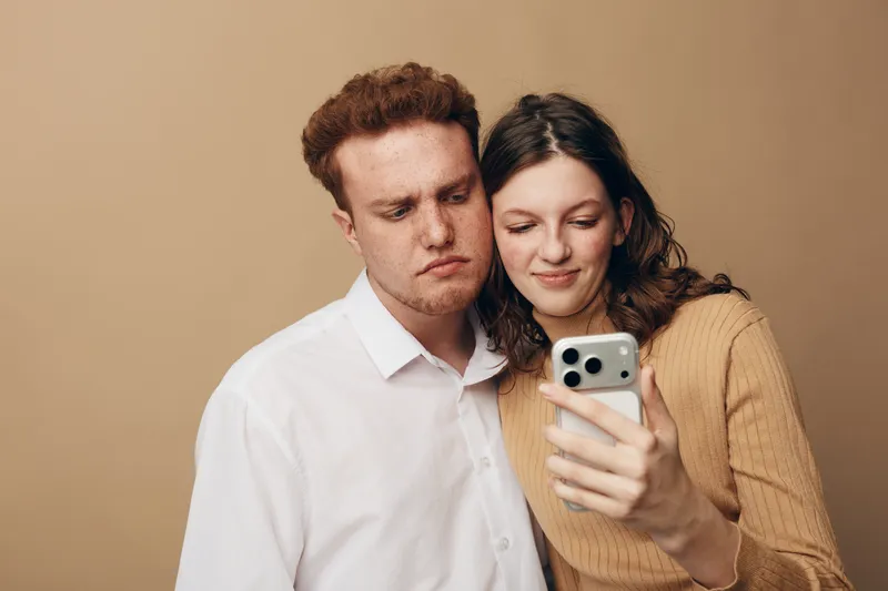Young couple looking at smartphone screen together with neutral expressions. Casual clothing and beige background create a simple, intimate moment of shared focus and connection.