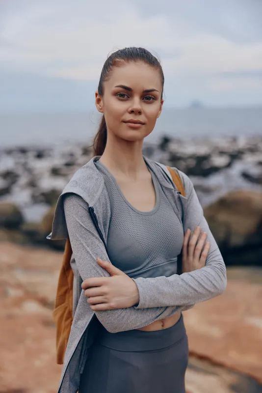 Young athletic woman in sportswear, standing confidently on rocky beach, with ocean in the background, conveying a sense of empowerment and health