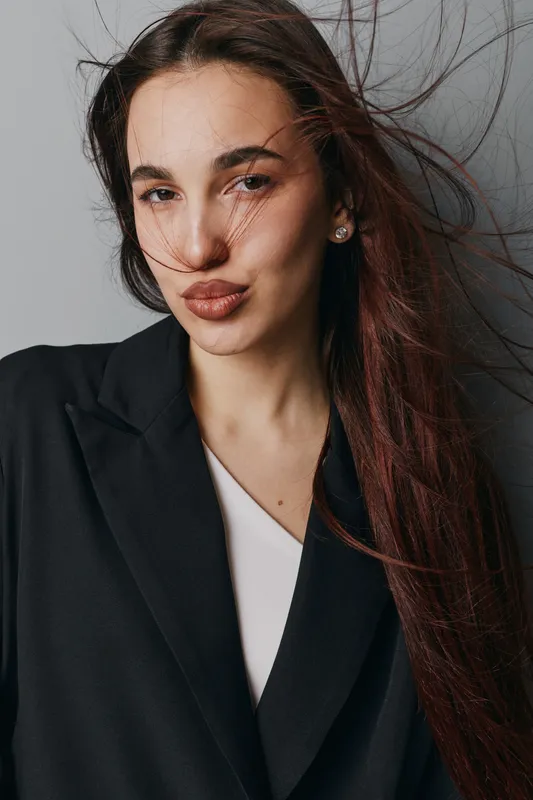 Woman with long brown hair blowing in the wind wearing black blazer and white top. Portrait of young female with natural makeup and confident expression on neutral background.