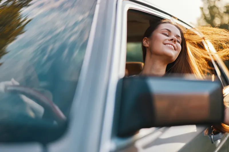 Young woman enjoying a joyful moment with her hair blowing in the wind, sitting in a car, surrounded by nature on a sunny day.