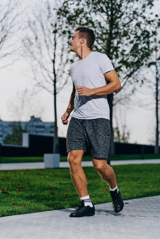 Спортивный мужчина в шортах и футболке готовится к пробежке на природе, в парке, возле деревьев, на улице