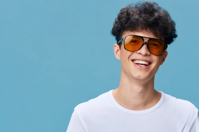 Smiling young man with curly hair wearing yellow tinted sunglasses and a white t-shirt on a blue background, expressing happiness and casual style in bright studio light.