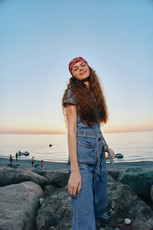 Smiling woman with long curly hair wearing denim outfit and red bandana stands on rocky beach during sunset with calm sea and clear sky in the background.