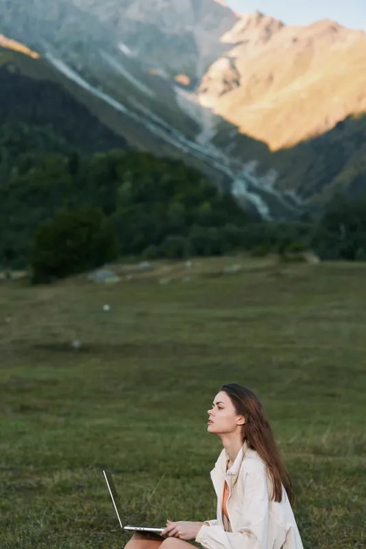 A calm woman sits on grass with a laptop in a mountainous meadow. She gazes upward, concentrating on work, creating a serene and focused outdoor workspace.