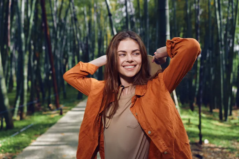 Woman smiling portrait in autumn park wearing an orange jacket on a tree-lined pathway. Outdoor lifestyle scene with relaxed young adult, natural light, casual fashion and joyful expression.