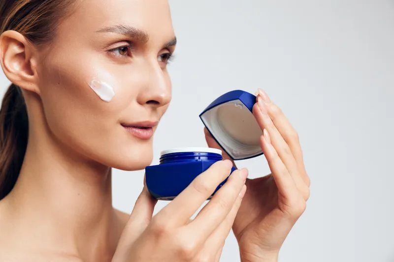 Young woman applies moisturizing cream on her face holding open jar with white lotion close to skin, demonstrating beauty skincare routine on neutral background.