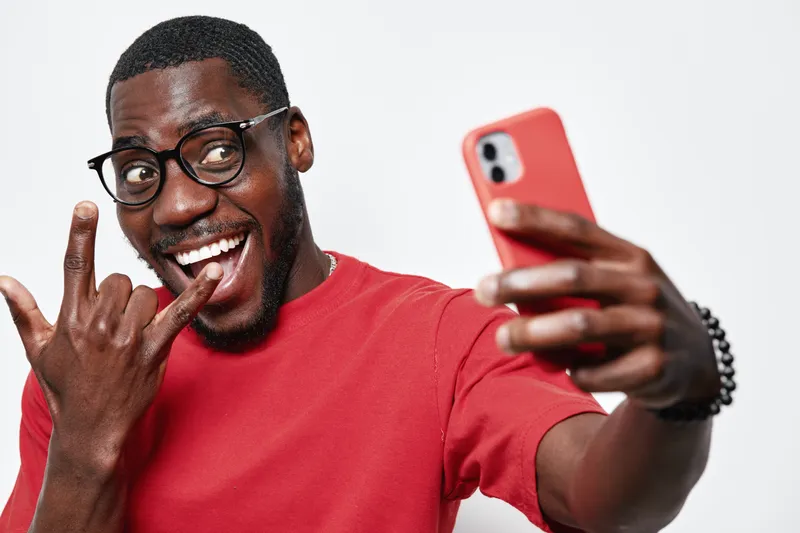 Black man with dark skin and glasses taking selfie with smartphone, smiling and making rock sign with hand on white background. Casual red shirt and happy expression.