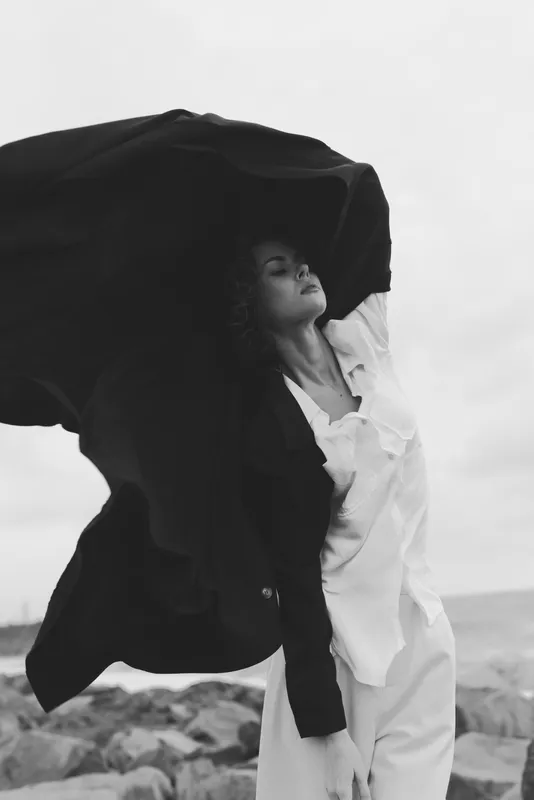 Woman in flowing cape raises fabric against wind on a rocky beach, fashion photo with dramatic movement, monochrome tones, serene and powerful mood, outdoor fashion imagery with natural light and