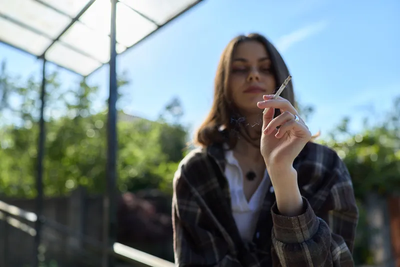 A relaxed woman in a plaid shirt stands outdoors, holding a cigarette toward the camera. Bright daylight, shallow depth of field, and casual vibe create a lifestyle portrait