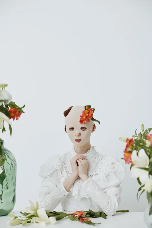 A surreal portrait of a model with a smooth mask and floral accents, wearing a white lace dress, evoking emotions of mystery and beauty in a minimalistic setting