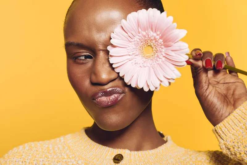 Young black woman with dark skin holds pink flower covering one eye, wearing yellow sweater on yellow background. Concept of beauty, skincare, nature, and self-expression.