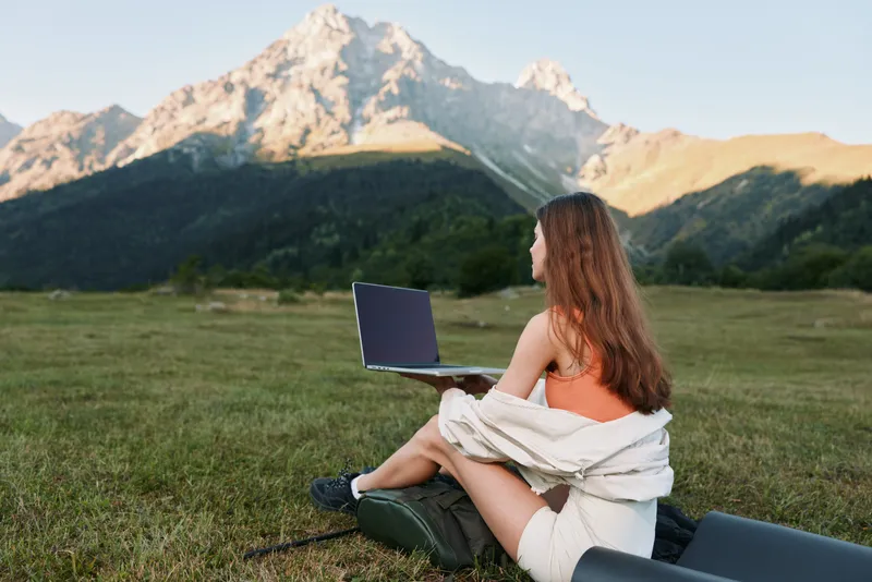 A relaxed woman sits on a blanket with a laptop in a scenic meadow. She wears casual attire and looks focused, enjoying productive remote work in a peaceful outdoor setting.