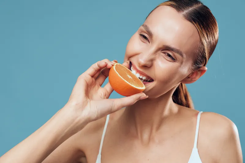 Young woman with brunette hair smiles joyfully while holding a half orange near her mouth against a plain blue background in a bright white top.