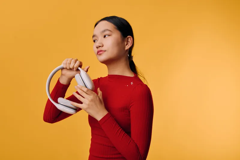 Young woman in red sweater holding white headphones looks away on yellow background with thoughtful expression. Studio portrait with casual clothing and modern accessory in hand.