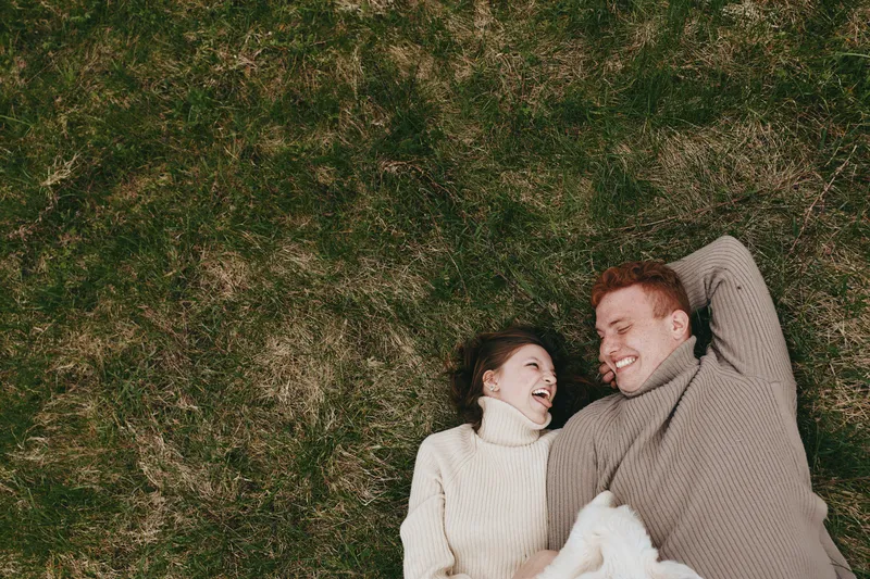 A couple lying on grass, smiling and relaxed, enjoying outdoor leisure together in cozy sweaters, sharing a warm moment in a natural park setting.