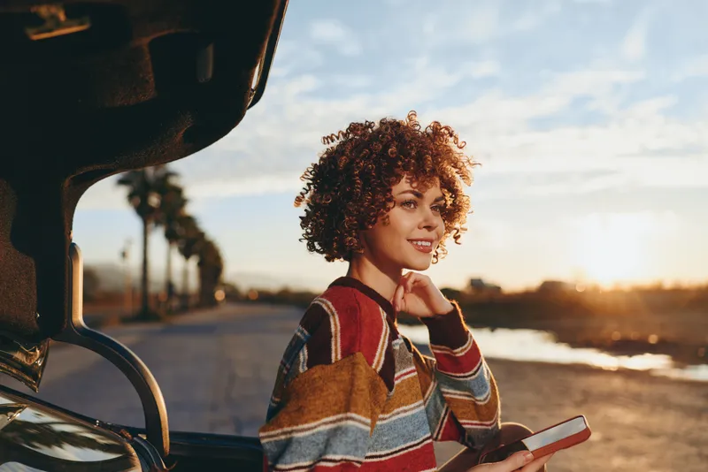 Woman lifestyle outdoors, car near beach, smiling with curly hair, wearing striped sweater, holding smartphone, golden sunset light, warm breeze, candid travel moment, soft natural colors
