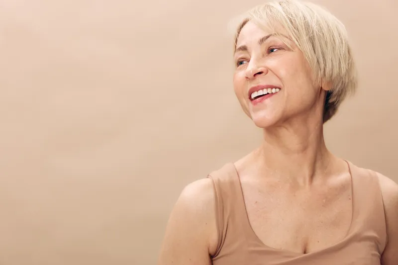 Smiling mature woman with short blonde hair looks away in beige tank top, isolated on soft light brown background. Concept of happiness, confidence, natural beauty, and positive aging.