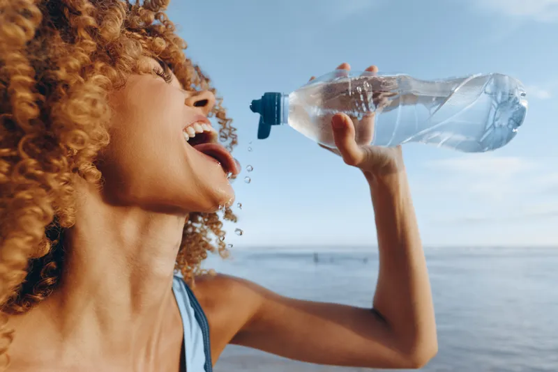 A young woman with curly hair drinks water from a plastic bottle near the sea under a clear blue sky. Concept for hydration, summer, and healthy lifestyle outdoors.