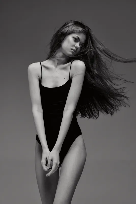 Black and white portrait of a slender woman with flowing hair wearing a black bodysuit expressing calm emotion in a minimalistic studio setting.
