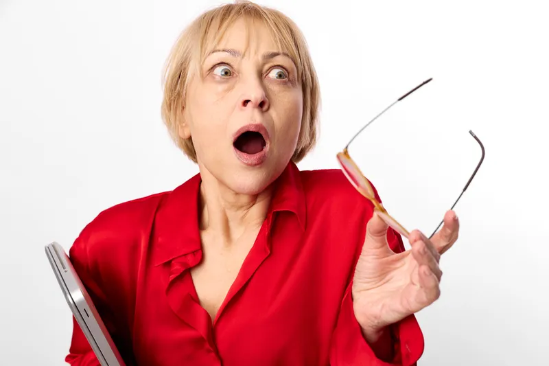 Elderly woman, surprised, shocked expression, holding glasses with a tablet, red blouse, wide open mouth, studio white background, bright lighting, closeup portrait, copy space