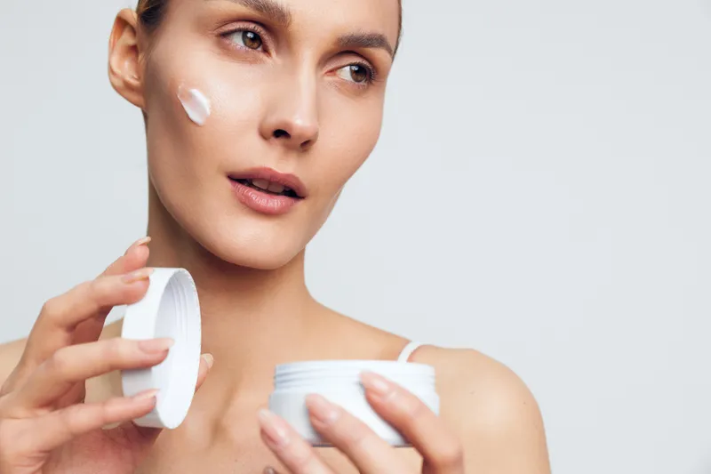Woman skincare, face cream applying, beauty spa hygiene, young woman holds cotton pad near cheek, calm expression, smooth facial skin, studio close up on white background.