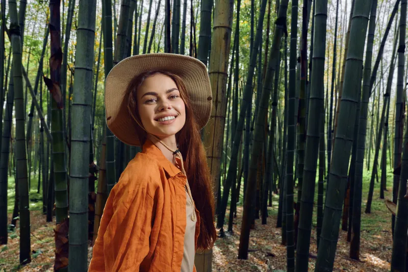 Woman with a smile in a bamboo forest wearing a straw hat and orange jacket, portrait in nature outdoor setting. Young traveler enjoying greenery, sunlight and peaceful mood.