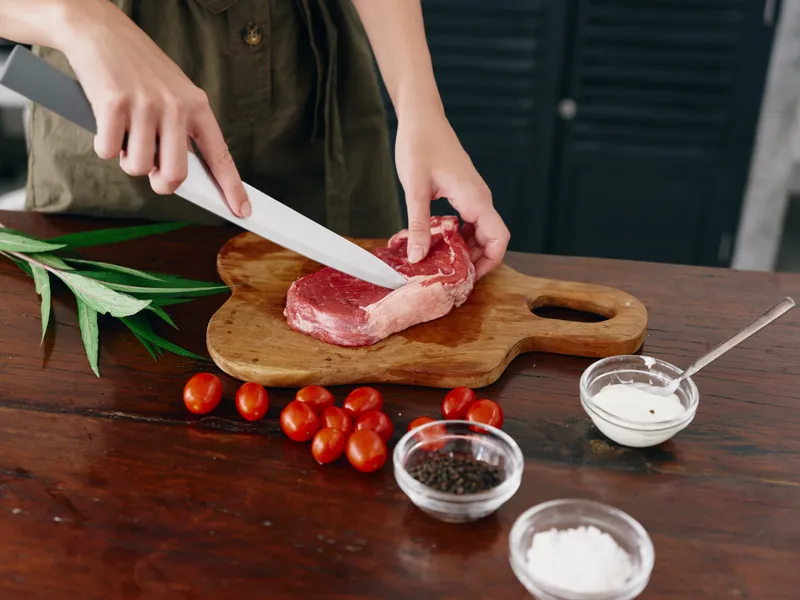 Женщина с ножом в руке нарезает свежее мясо для стейка для запекания на кухне, на столе соль, перец и другие специи, красные помидоры черри и зелень, приготовление ужина