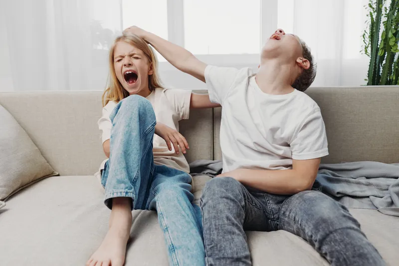 Two children sitting on a sofa playfully yelling and pulling each other's hair in a bright living room. Concept of sibling rivalry, childhood emotions, and family dynamics captured naturally.