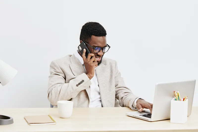 Professional man with dark skin, wearing glasses and beige blazer, talking on the phone while working on a laptop in a modern office space. Business, communication, technology, workspace, people conce