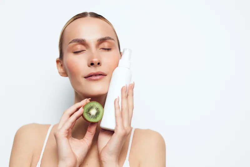 A young woman with clear skin holds a white cosmetic bottle and a halved kiwi fruit near her face. She has closed eyes and a serene expression against a plain white background.