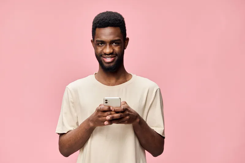 Smiling young African man holding smartphone in casual beige t shirt against pink background, reflecting happiness and modern lifestyle