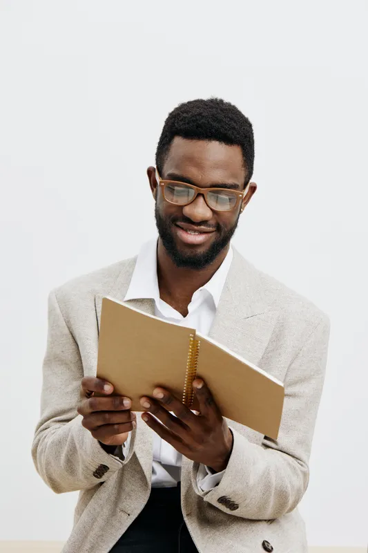 Smiling man with glasses reading a notebook, dressed in a beige blazer and white shirt, isolated on white background. Casual, intellectual, professional, relaxed, stylish. People lifestyle concept.