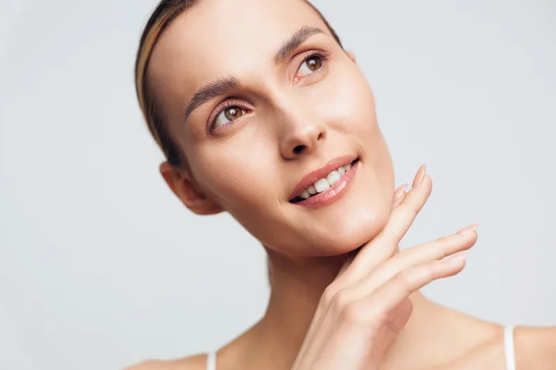 Young woman with clear skin and natural makeup gently touches her face and looks away with soft smile, isolated on light background, beauty and skincare concept.
