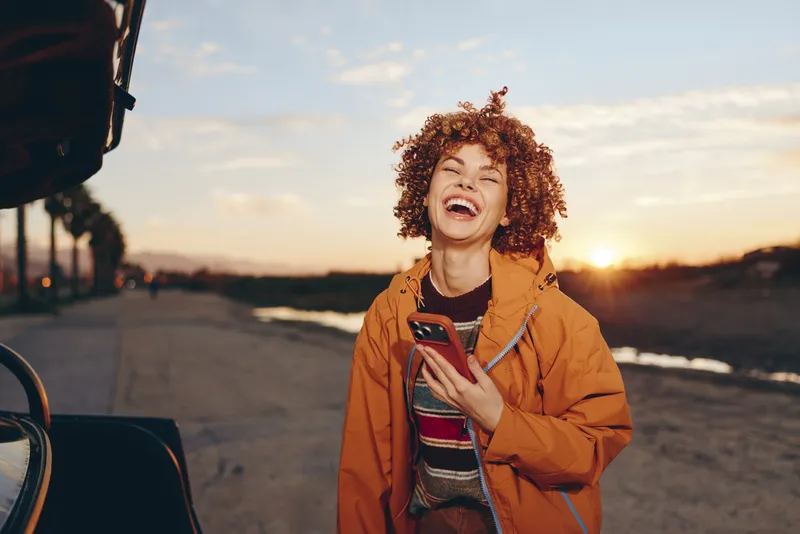 Woman lifestyle outdoors at sunset, casual young woman laughs with a phone in hand beside a car on the road, warm golden light, travel moment, candid smile
