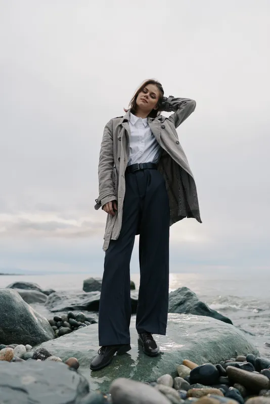 Young woman in casual outfit stands on rocky shore under cloudy sky. She wears long coat and wide pants, posing with hand on head near sea. Concept of fashion and nature.
