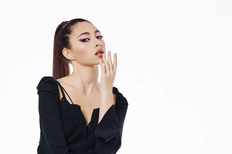 Stylish woman in black fashion outfit poses with confident expression and hand near face. She has sleek hair and dramatic makeup on white background. Creative beauty concept.